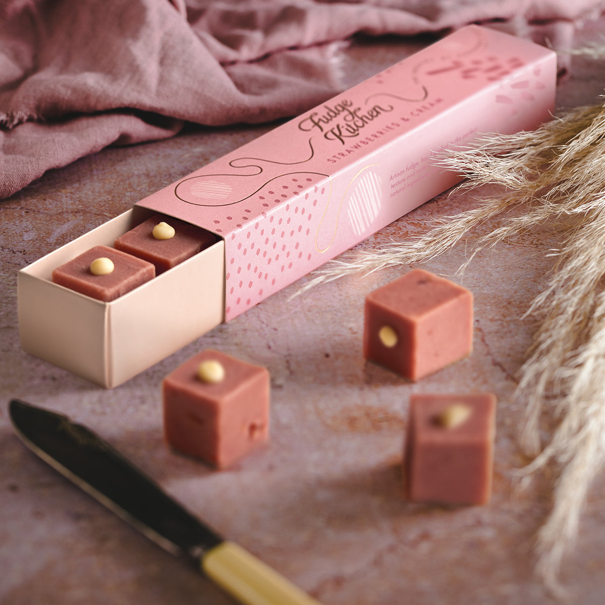 Pink fudge selection box with pink cubes of strawberries & cream fudge, displayed on a textured surface.