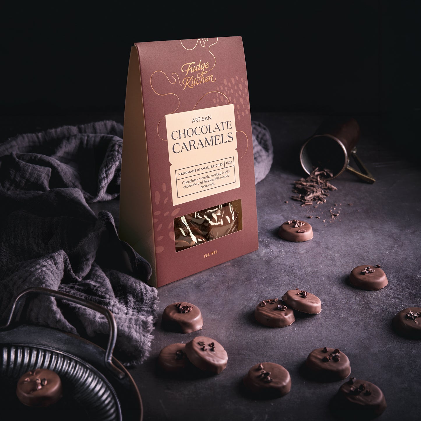 Box of Fudge Kitchen Artisan Chocolate Caramels on a dark surface with caramels around on a table with a grey cloth.