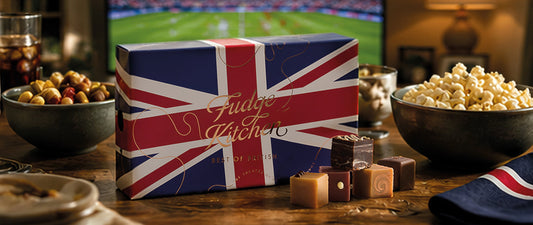Fudge Kitchen fudge selection box with Union Jack design on a table with popcorn and snacks, TV in the background.