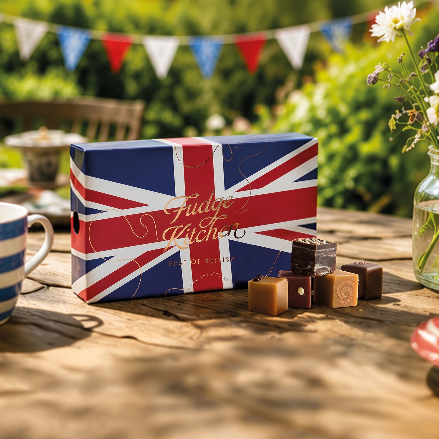 Fudge Kitchen Best of British Fudge Selection Box with a Union Jack flag design displayed on a wooden picnic table with snacks and drinks.