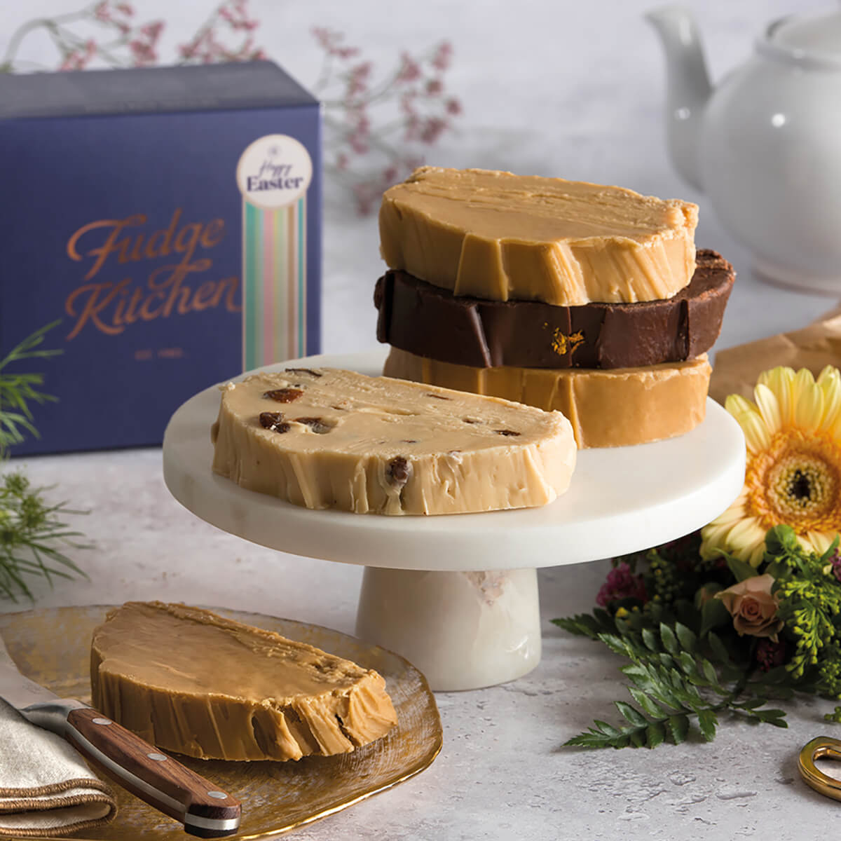 Vegan Easter fudge on a cake stand surrounded by flowers, with a blurred gift box in the background – a delightful and festive vegan treat for Easter.