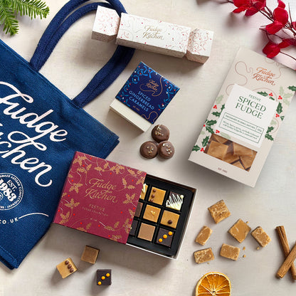 Fudge Kitchen Christmas gift set arranged on a light neutral surface with a navy tote bag, assorted fudges, a box of spiced fudge, a small box of spiced caramels, and decorative holiday props.