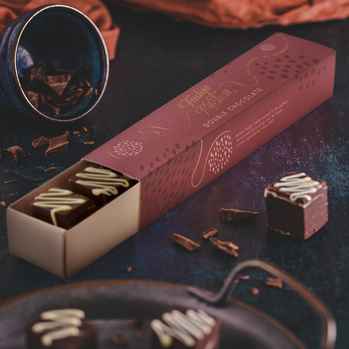 A box of double chocolate fudge next to two fudge cubes on a dark table, with a decorative background.