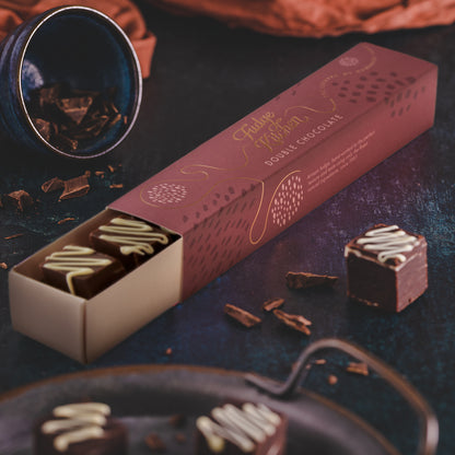 A box of double chocolate fudge next to two fudge cubes on a dark table, with a decorative background.