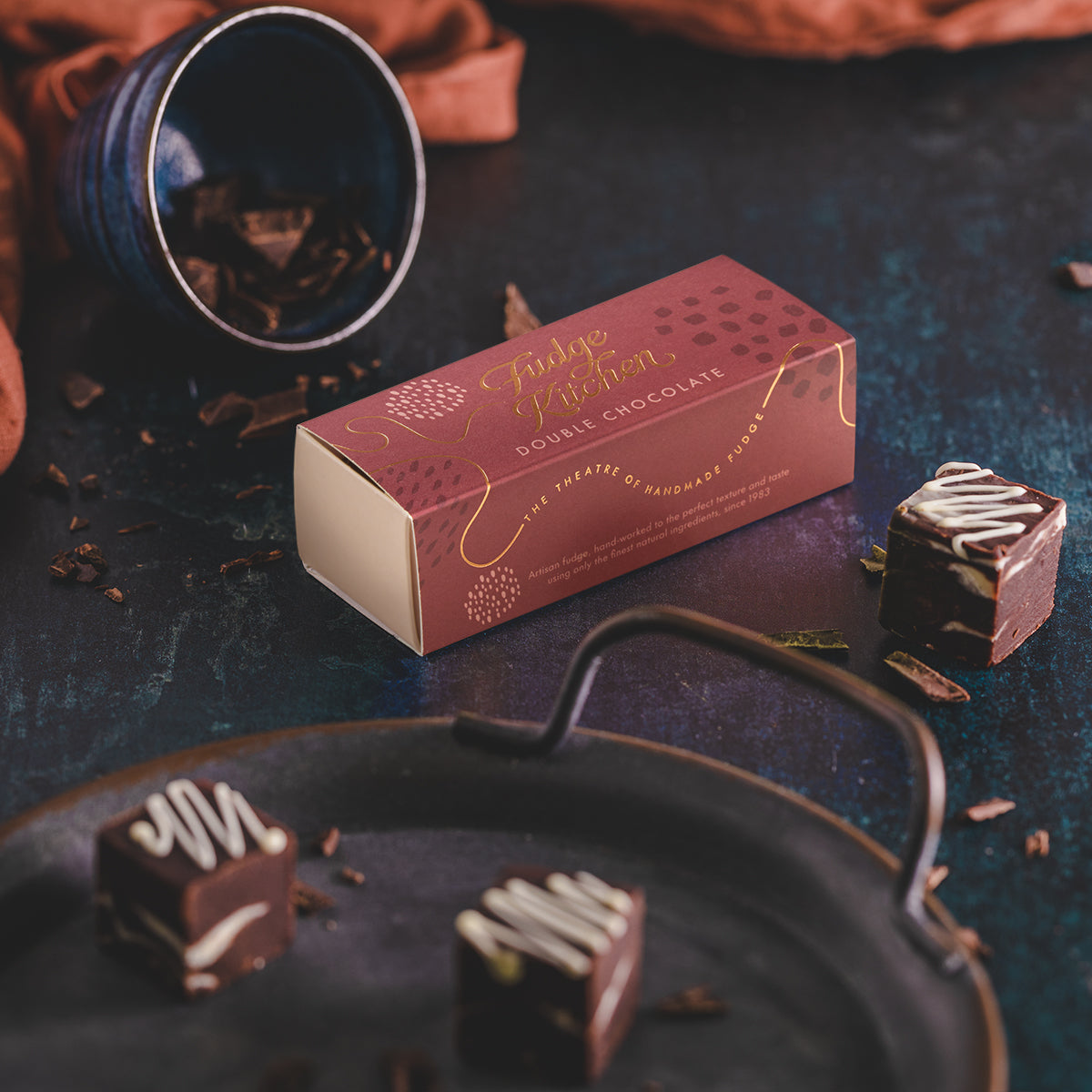 A box of double chocolate fudge with white chocolate swirls, placed on a dark surface with loose chocolate pieces around. The box is adorned with a chocolate-themed design.