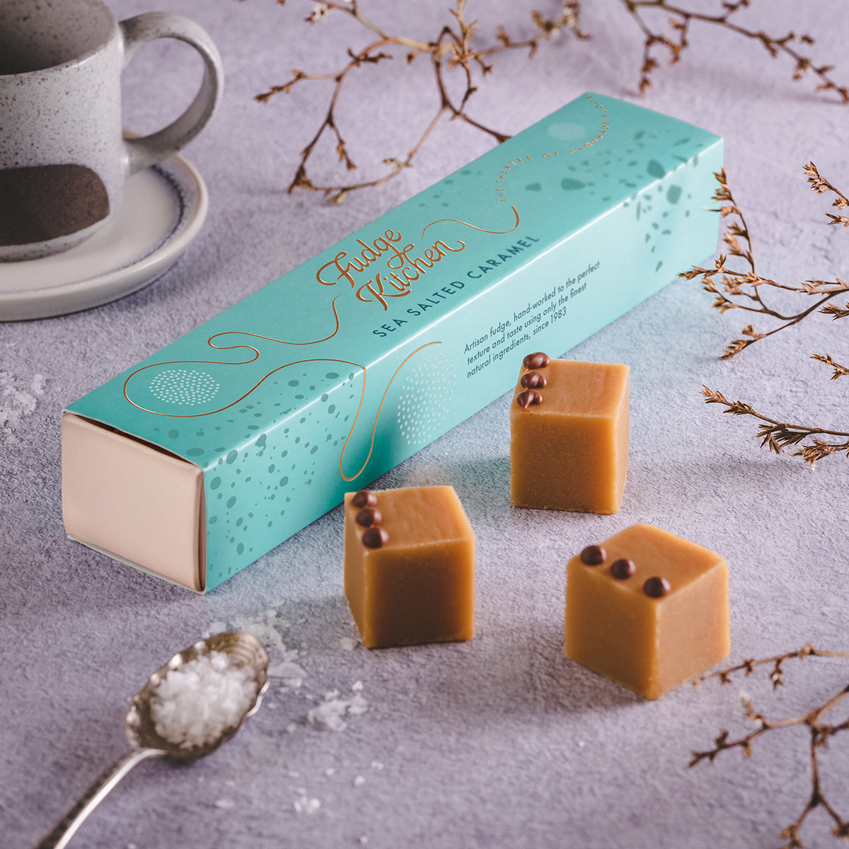A box of salted caramel fudge next to three fudge cubes on a grey table, with a decorative background.