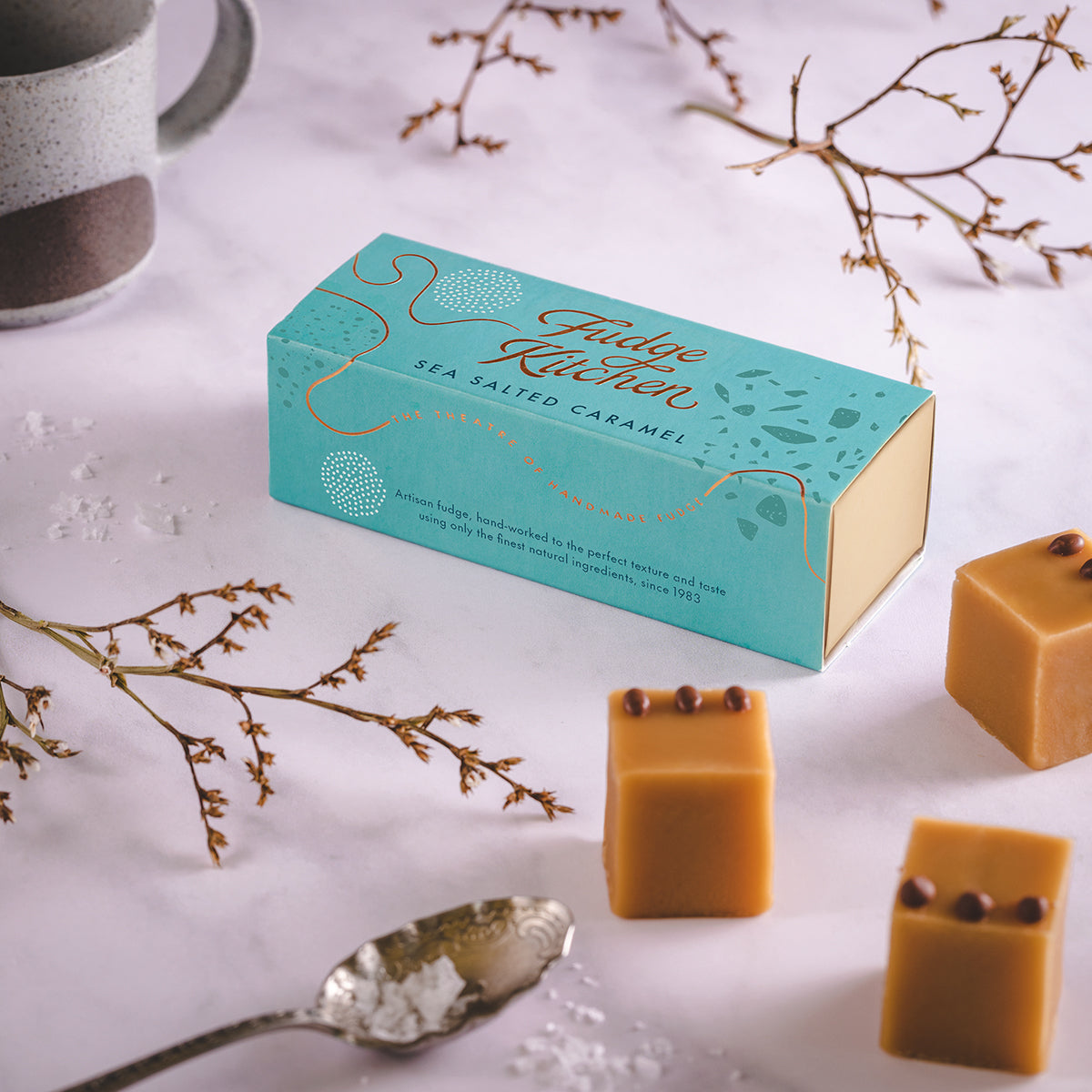 A box of salted caramel fudge next to three fudge cubes on a grey table, with a decorative background.