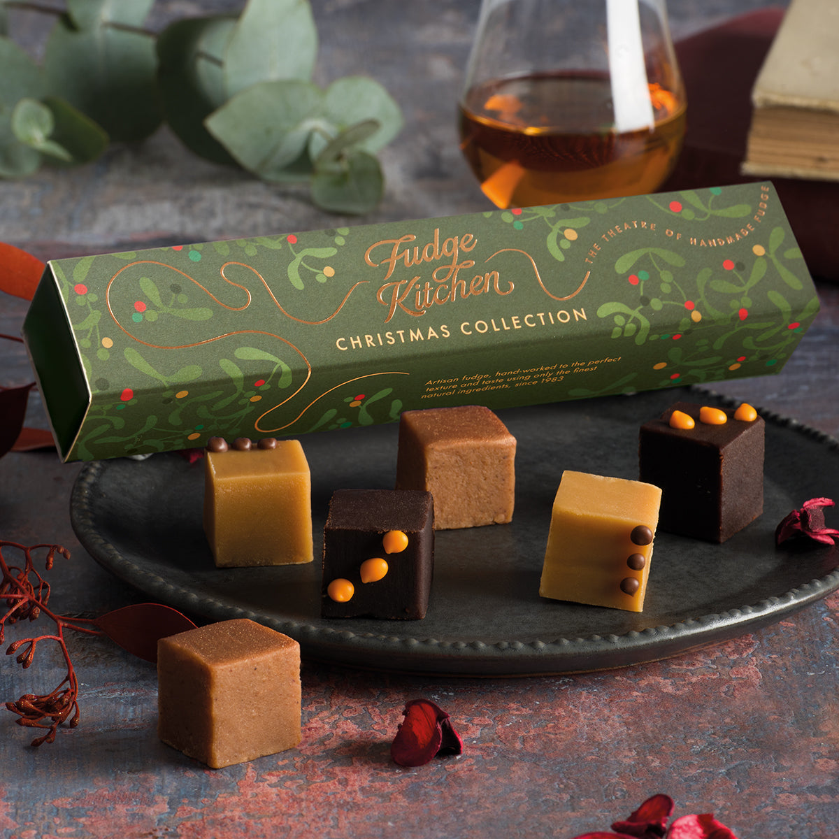 A box of christmas fudge next to fudge cubes on a surface, with a decorative background.