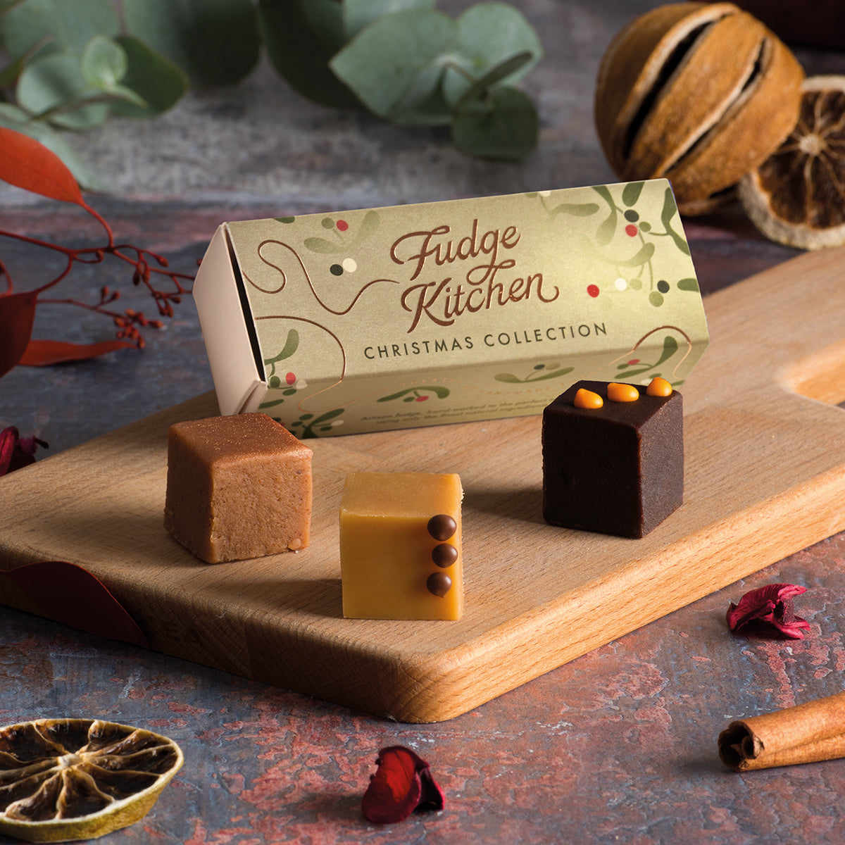 A box of christmas fudge next to three fudge cubes on a wooden board, with a decorative festive background.