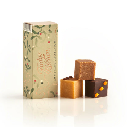 Three pieces of festive fudges stacked in front of the long Fudge Kitchen christmas gift box on a white background