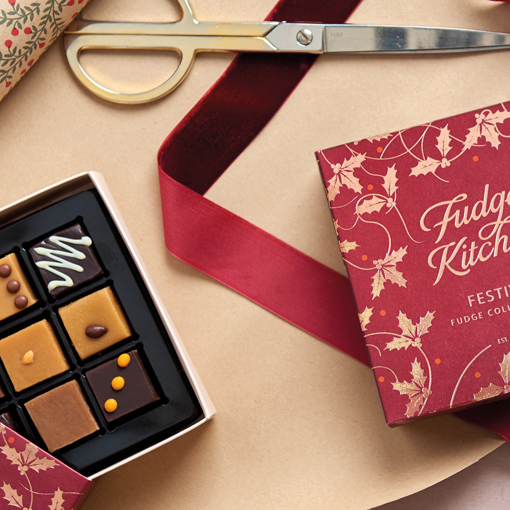 An open box of assorted fudge pieces beside a red gift box with gold holly leaf illustrations, arranged on a kraft paper background with gold scissors and a red ribbon.