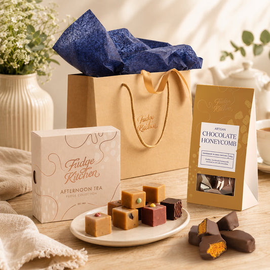 A gourmet fudge gift set from Fudge Kitchen on a wooden table. The set includes a kraft paper gift bag with blue tissue paper, a box of afternoon tea fudge, a package of artisanal chocolate honeycomb, and several decorated fudge squares on a small white plate.