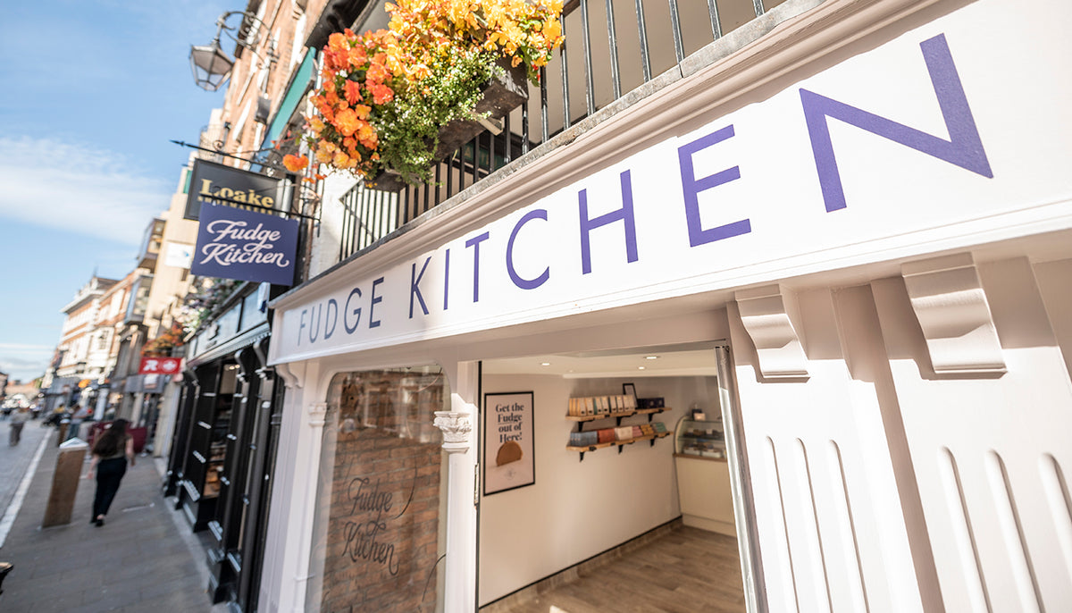 Fudge Kitchen storefront on a city street, with large indigoe lettering on a white facade, flower boxes hanging from the balcony, and a visible interior display.