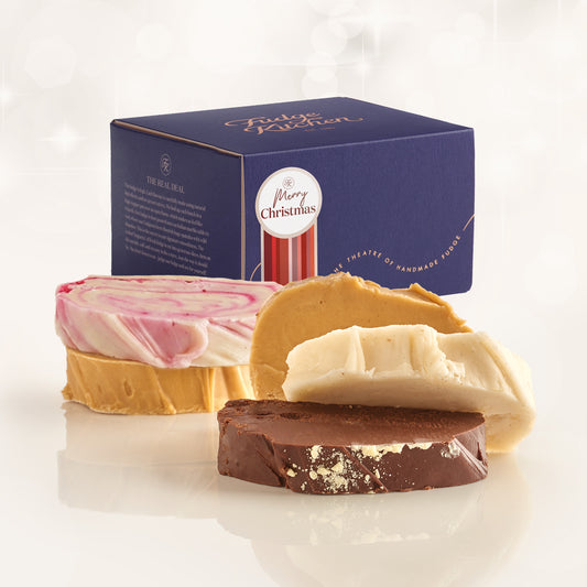 Four artisanal fudge slices in pink, caramel, white, and dark chocolate, arranged in front of a dark blue 'Fudge Kitchen' Christmas gift box on a reflective white surface.