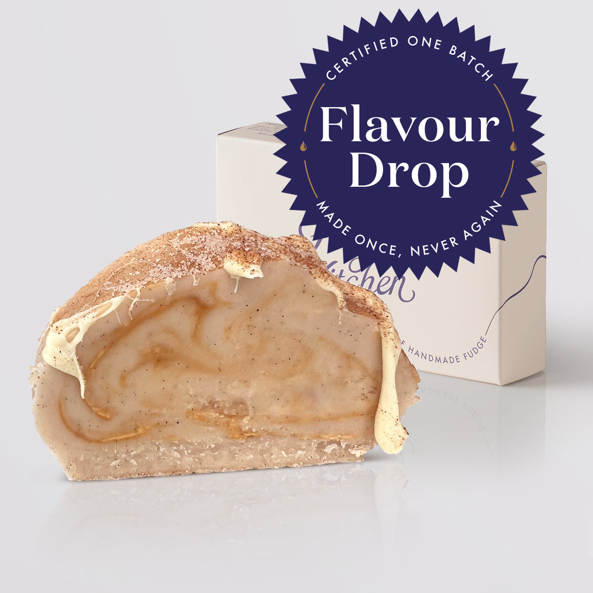 A single slice of cinnamon bun fudge show its creamy caramel swirl interior, placed in front of a white box with a large blue badge reading 'Flavour Drop' on a light gray studio background.