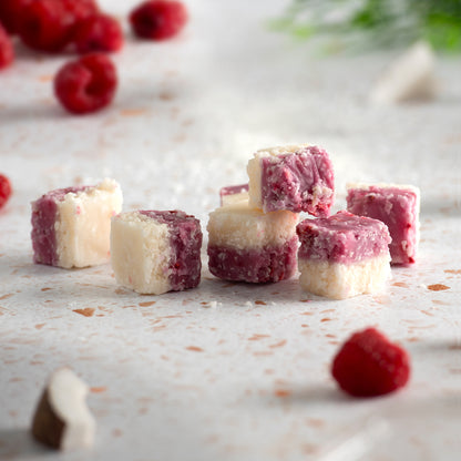 Raspberry coconut ice pieces on a light surface with raspberries in the background.