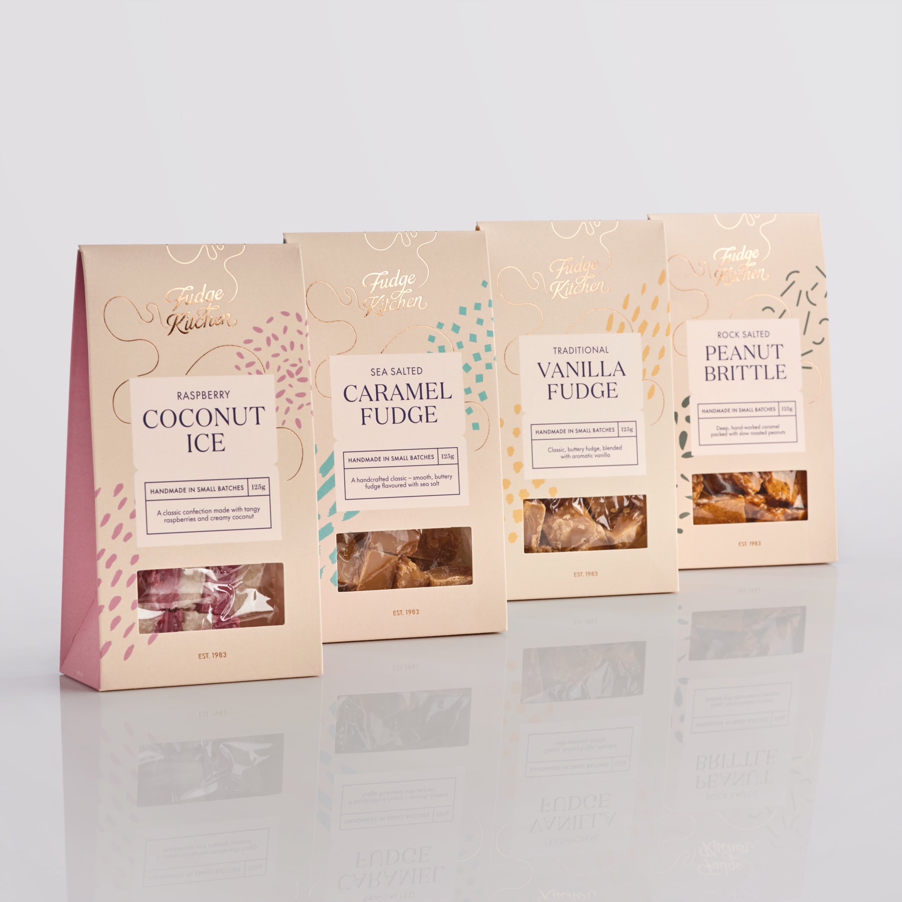 Four packages of Fudge Kitchen luxury confectionery on a reflective surface with a light background