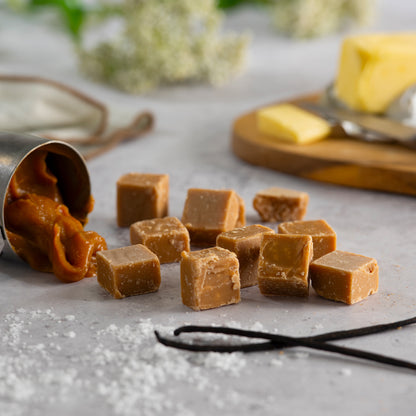 Traditional vanilla fudge pieces on a gray surface with a container of caramel and a wooden board with golden butter in the background.