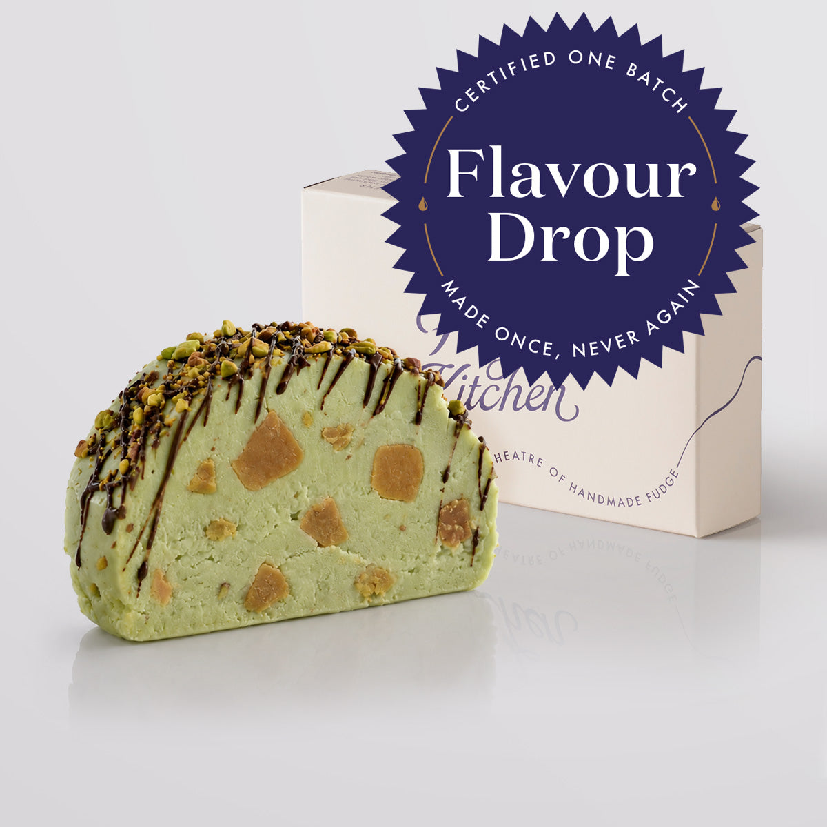 A slice of dubai pistachio chocolate fudge with chocolate drizzle and chopped nuts, shown in front of a gift box and a blue circular 'Flavour Drop' badge on a light grey background.