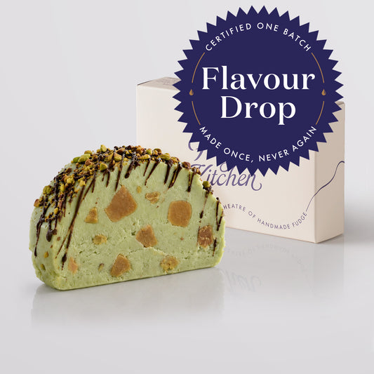 A slice of dubai pistachio chocolate fudge with chocolate drizzle and chopped nuts, shown in front of a gift box and a blue circular 'Flavour Drop' badge on a light grey background.