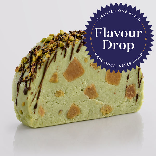 A single pistachio fudge sliced with embedded salted caramel fudge chunks, topped with chopped pistachio and dark chocolate drizzle, on a light gray background with a badge overlay containing the text 