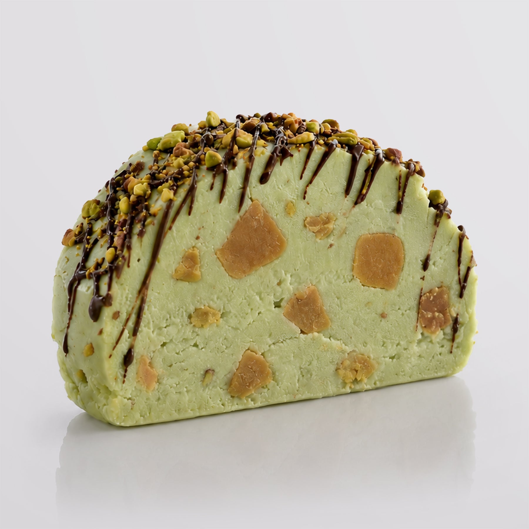 A single pistachio fudge sliced with embedded salted caramel fudge chunks, topped with chopped pistachio and dark chocolate drizzle, on a light gray background.