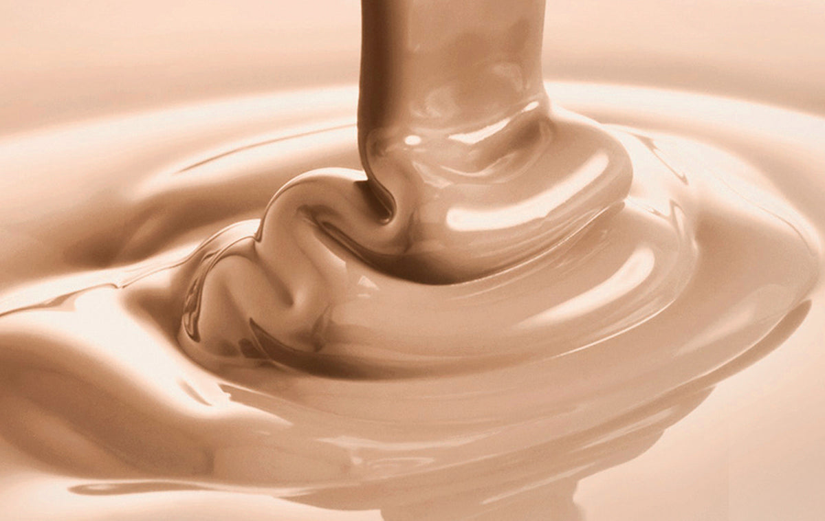 A close-up shot of a thick, glossy fudge, being poured in a smooth vertical stream into a shallow pool, creating ripples and splashes.