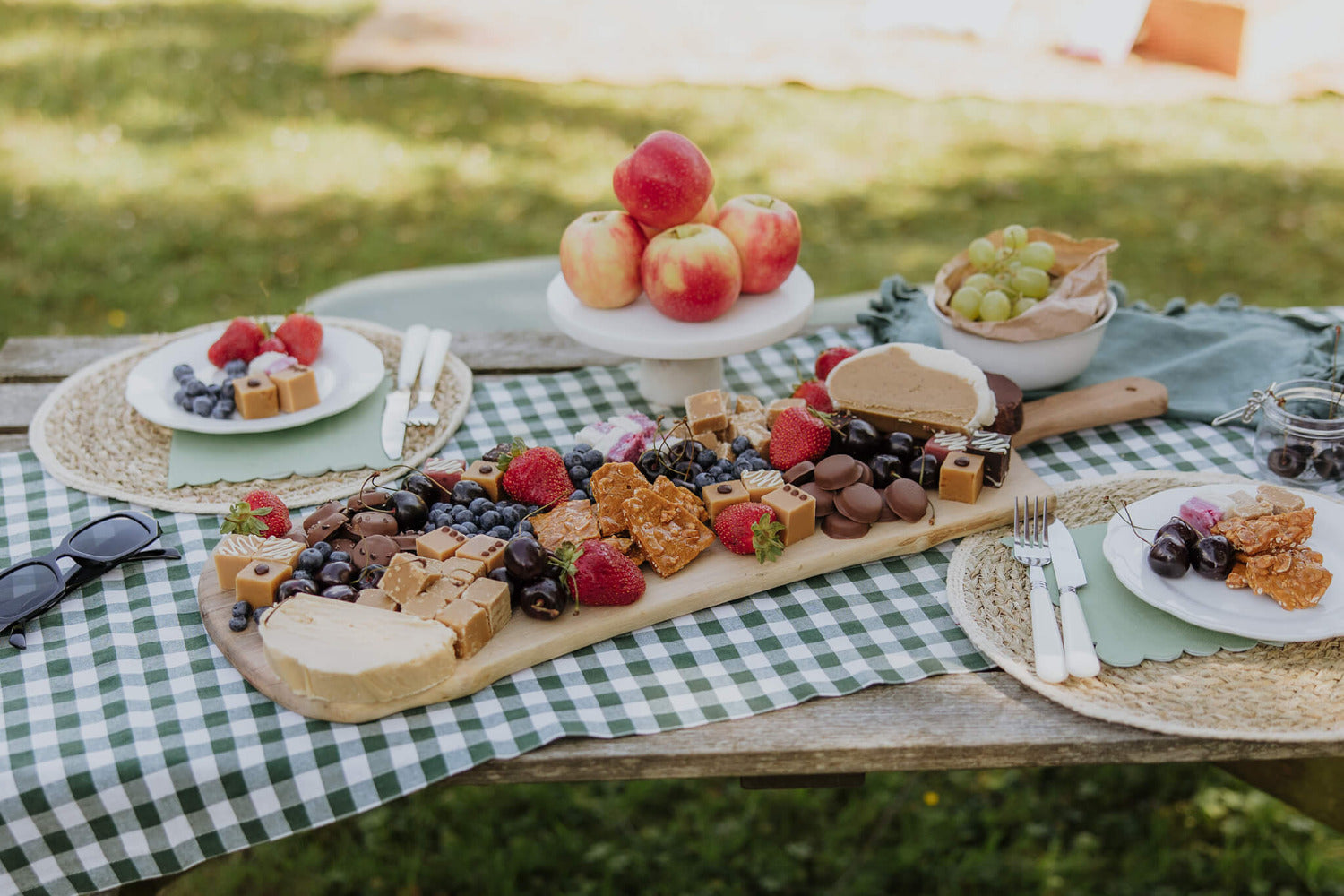 Picnic setup with a charcuterie board, fruits, fudge, chocolate caramels and peanut brittle on a checkered tablecloth outdoors.