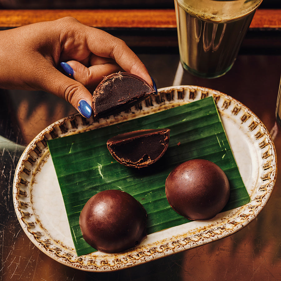 A hand with blue nail polish holds a chocolate-covered fudge dome above a plate with two round chocolate-covered spheres and a third broken half, arranged on a green banana leaf on a patterned ceramic plate.