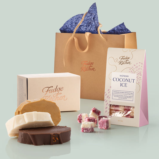 Fudge Kitchen vegan products including vegan fudge slices, a box of fudge, and a bag of raspberry coconut ice on a light green background displayed with a gold foiled, luxury gift bag behind.