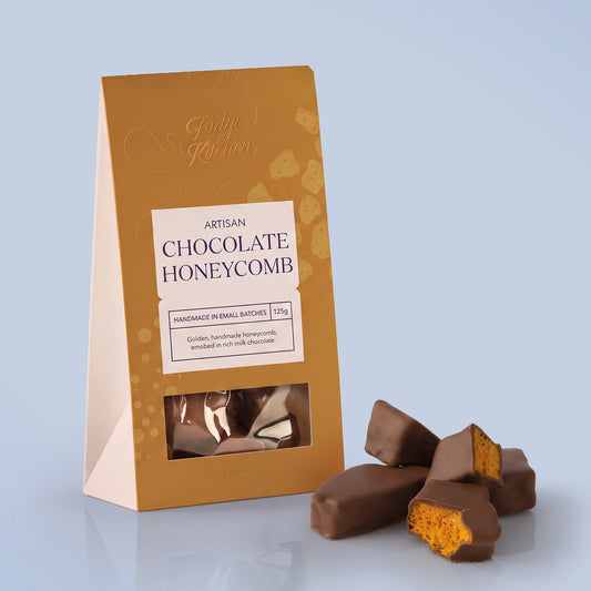 A gift pack of Fudge Kitchen Artisan Chocolate Honeycomb, shown next to several pieces of the chocolate covered honeycomb on a light blue background.