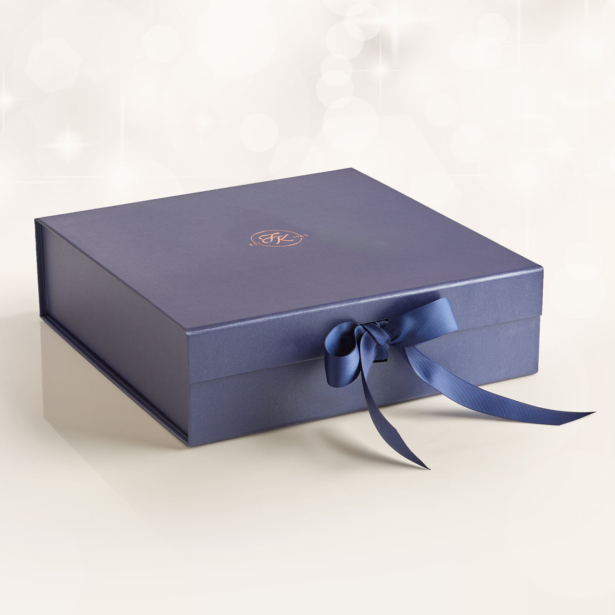 A dark blue rectangular solid, hamper gift box with a matching blue ribbon bow on the front and a small gold monogram logo on the lid, set against a soft white background with subtle bokeh light effects.