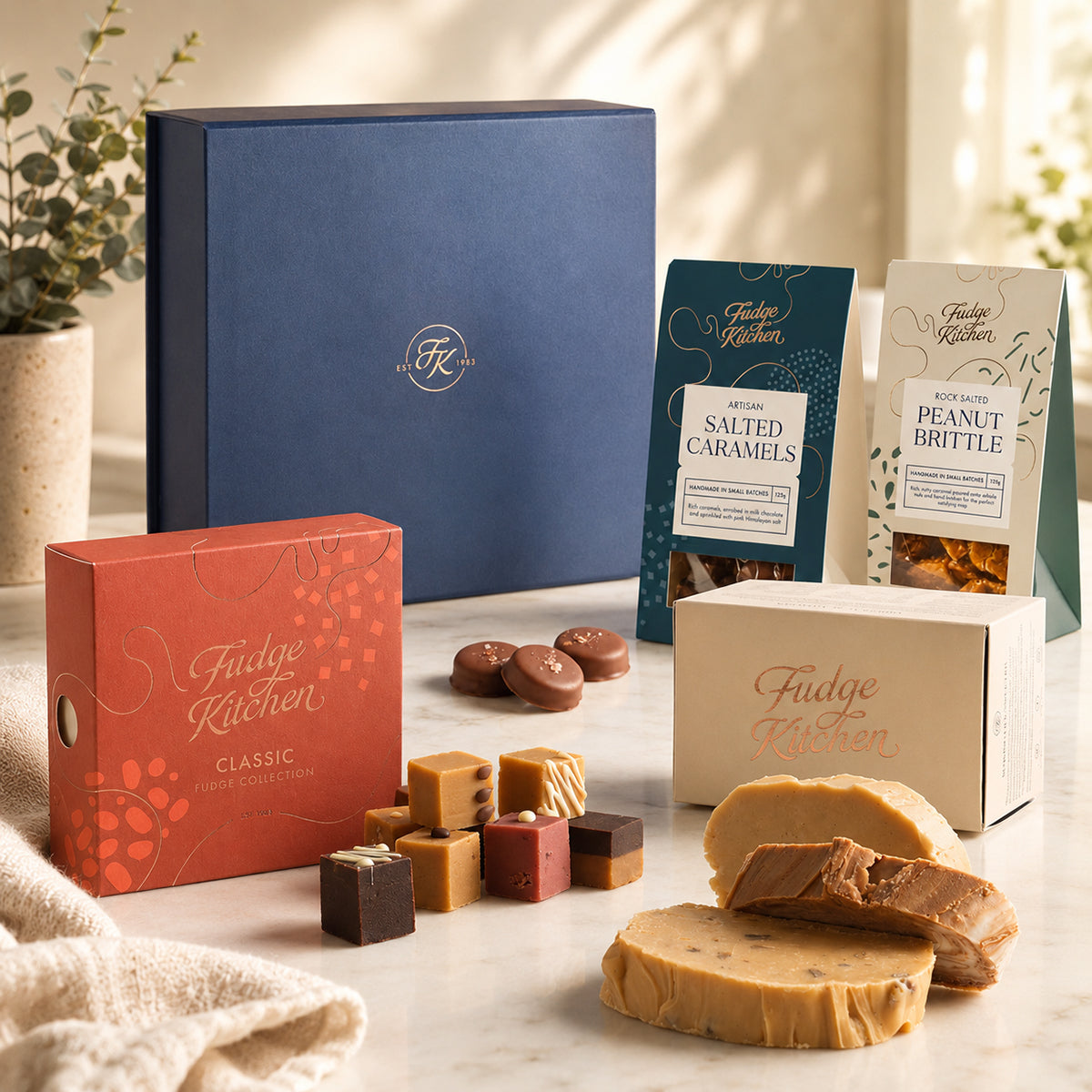 Fudge and luxury sweet gifts from Fudge Kitchen shown with packaging, including fudge, salted caramels, peanut brittle, and three slices of fudge on a table with a large premium hamper box displayed in the background.