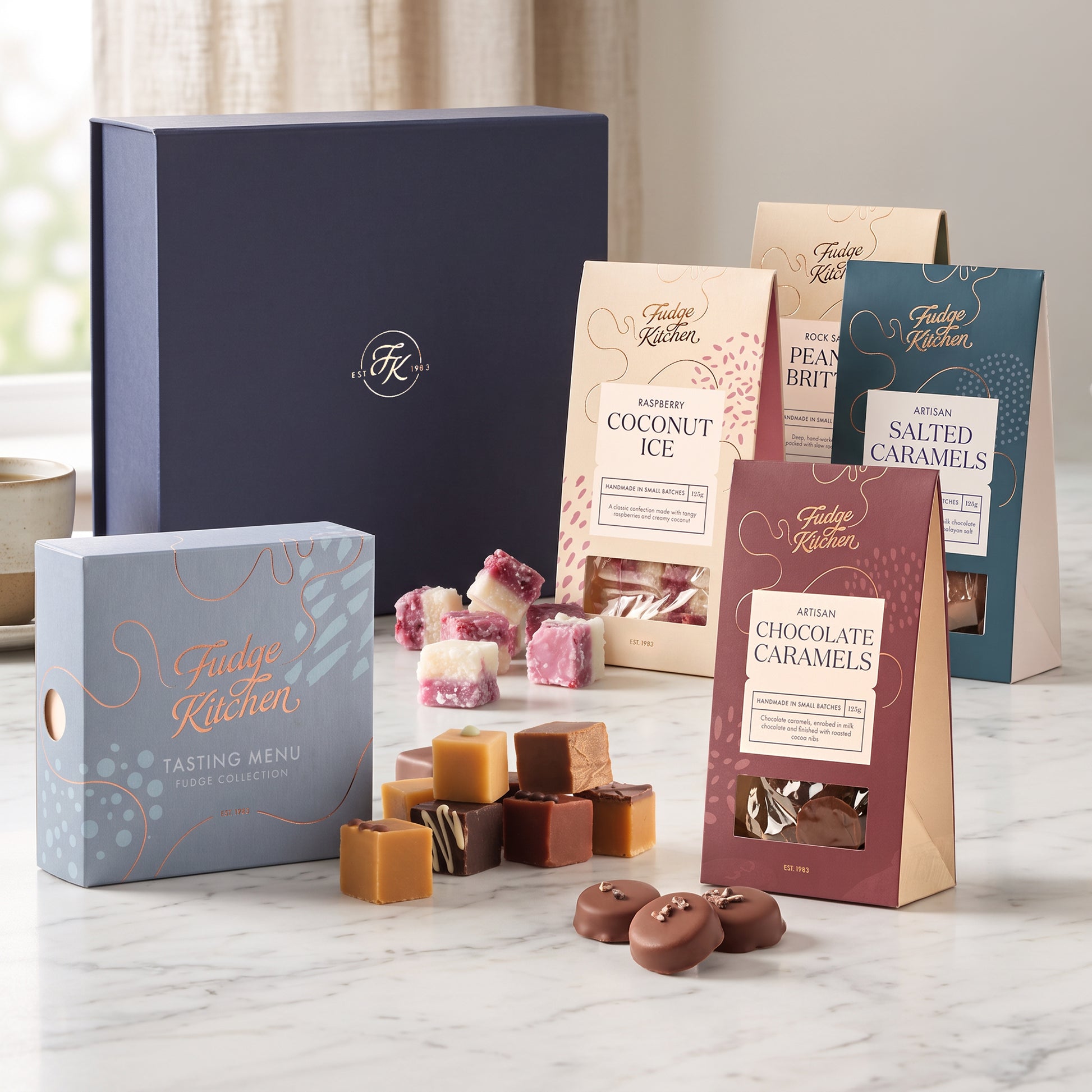 A curated assortment of Fudge Kitchen chocolates and packaged treats arranged on a light marble surface in front of a dark navy hamper gift box. Visible items include a blue box labeled Tasting Menu, several packs of coconut ice, salted caramels, and chocolate caramels, plus loose pieces of assorted caramels and fudge.