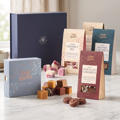 A curated assortment of Fudge Kitchen chocolates and packaged treats arranged on a light marble surface in front of a dark navy hamper gift box. Visible items include a blue box labeled Tasting Menu, several packs of coconut ice, salted caramels, and chocolate caramels, plus loose pieces of assorted caramels and fudge.