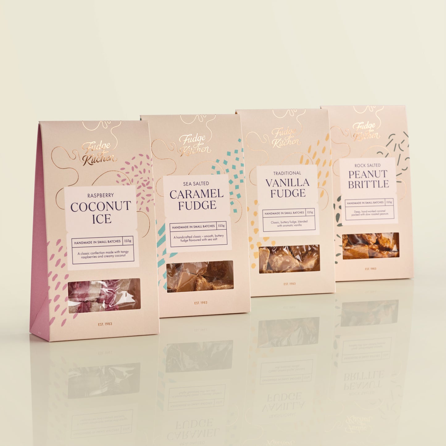 Four packages of Fudge Kitchen artisan confectionary gift packs on a reflective surface with a light background