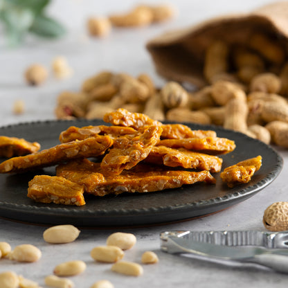 Peanut brittle on a black plate with peanuts scattered around on a light background