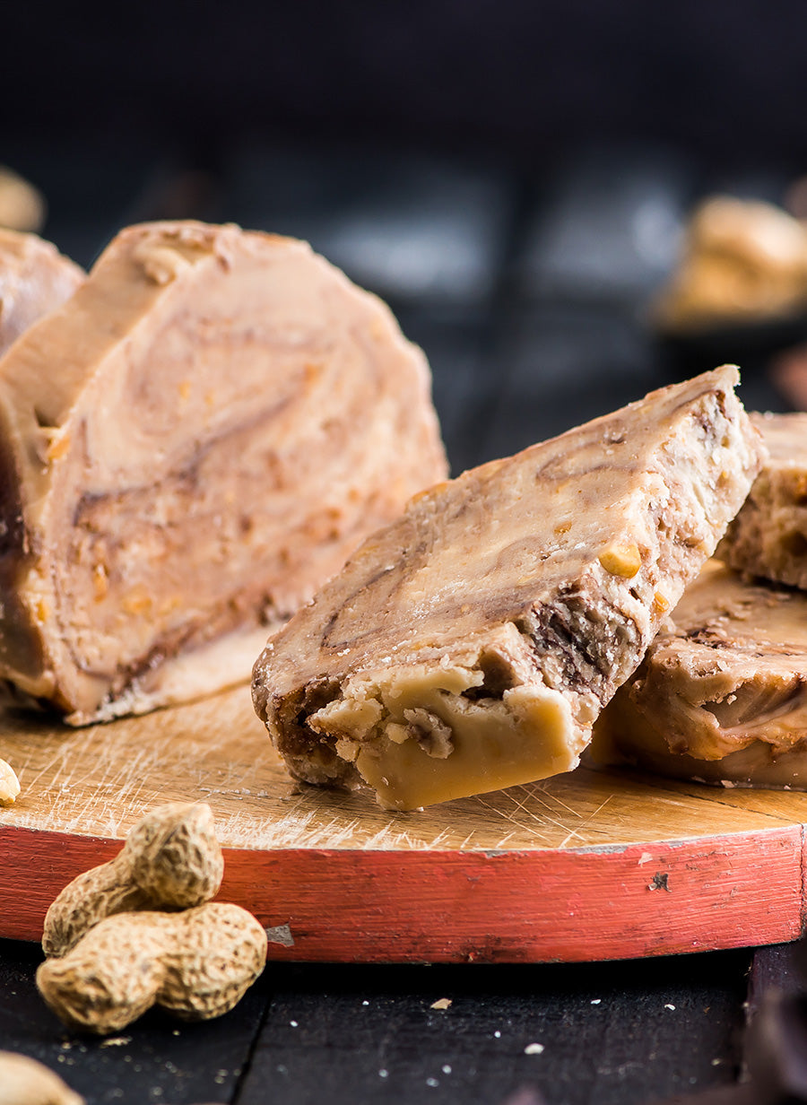 Slices of Peanut Butter Chocolate Swirl product displayed on a wooden cutting board with peanuts in the foreground, set against a dark background.