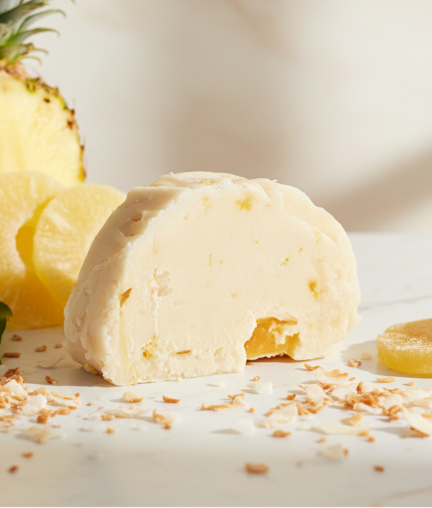 A slice of pina colada fudge on a marble table with a large pineapple behind it and two slices of pineapple in front of the fudge