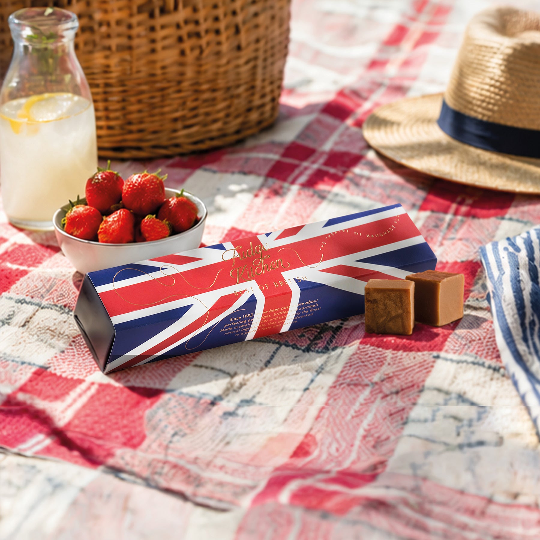 Picnic setup with strawberries, a Union Jack, Best of British Fudge Selection box, and a straw hat on a checkered tablecloth.