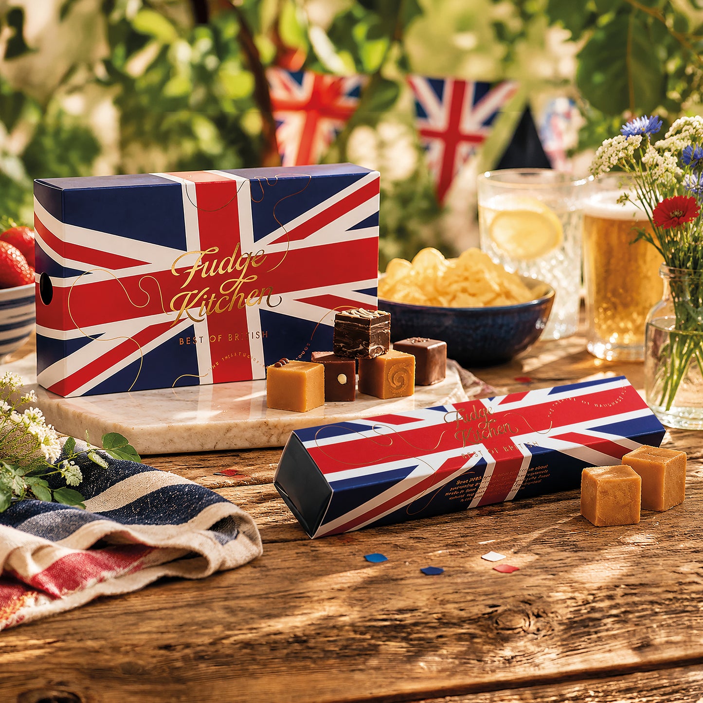 Fudge Kitchen Best of British fudge boxes with Union Jack design on a wooden table with snacks and drinks.