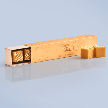 Yellow fudge packaging with 'Fudge Kitchen' branding, slightly open to reveal a piece of clotted cream fudge, with two pieces of artisan fudge in front of the long on a light blue background.