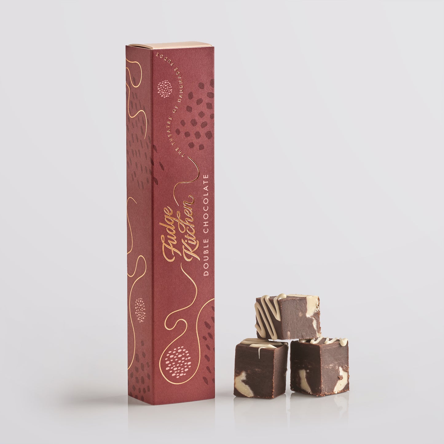 Three pieces of Fudge Kitchen double chocolate fudge stacked in front of a tall gift box on a light gray background.