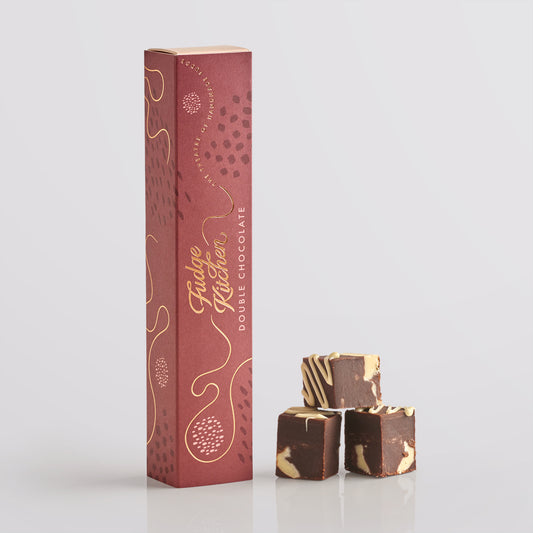Three pieces of Fudge Kitchen double chocolate fudge stacked in front of a tall gift box on a light gray background.