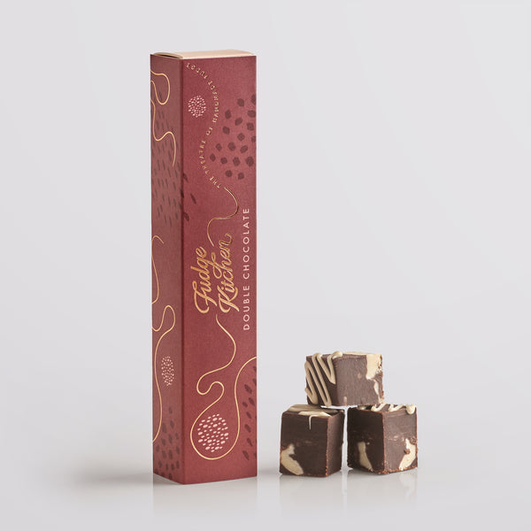 Three pieces of Fudge Kitchen double chocolate fudge stacked in front of a tall gift box on a light gray background.