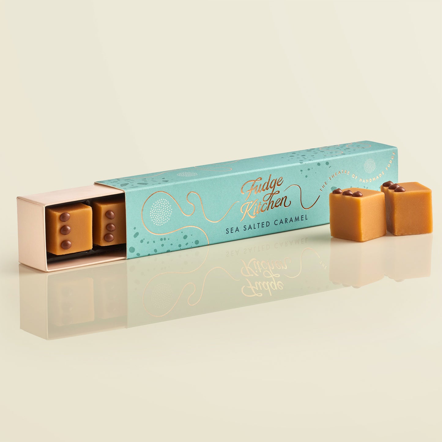 Teal fudge packaging with 'Fudge Kitchen' branding, slightly open to reveal a piece of salted caramel fudge, with two pieces of artisan fudge in front of the long on a beige background.