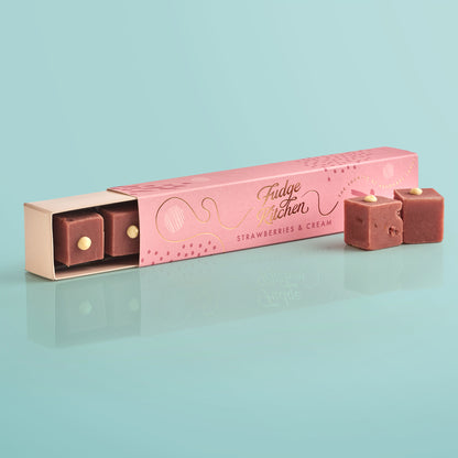 Pink fudge packaging with 'Fudge Kitchen' branding, slightly open to reveal a piece of strawberries & cream fudge, with two pieces of artisan fudges in front of the long on a light blue background.