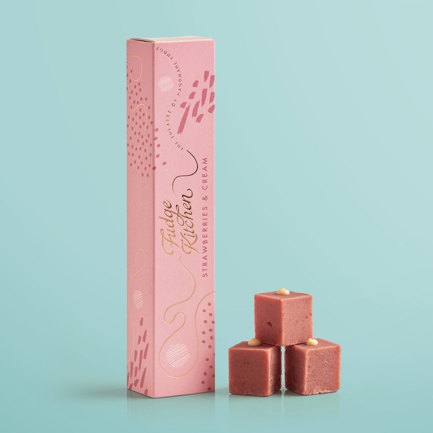 Three pieces of strawberries & cream festive fudges stacked in front of the long Fudge Kitchen gift box on a blue background.