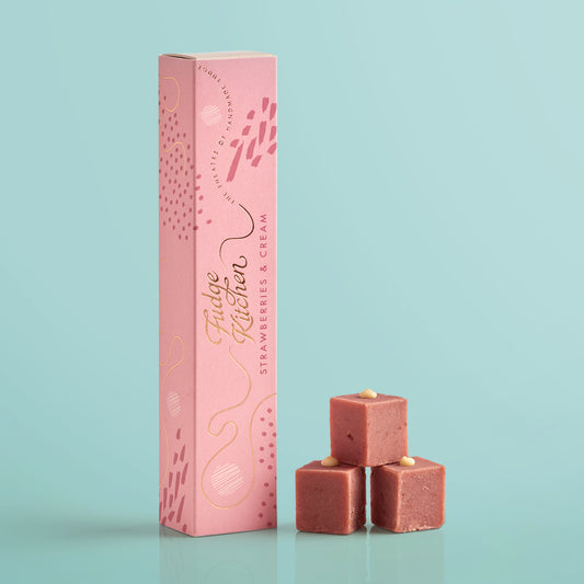 Three pieces of strawberries & cream festive fudges stacked in front of the long Fudge Kitchen gift box on a blue background.