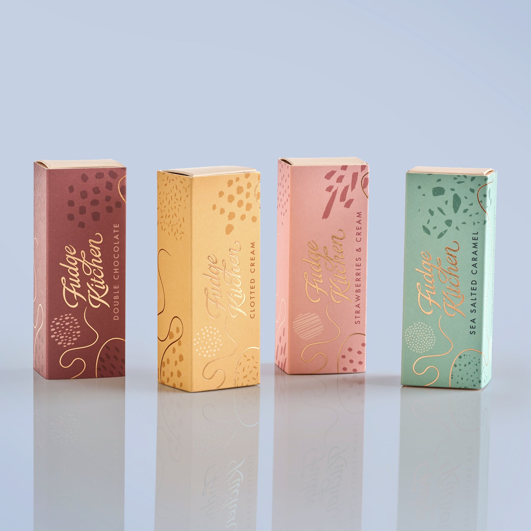 Four gift selection boxes of Fudge Kitchen products on a reflective surface with a light blue background
